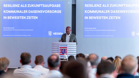 Der Minister steht auf einer Bühne und hält eine Rede - im Hintergrund einer Powerpoint mit dem Text "Resilienz als Zukunftsaufgabe kommunaler Daseinsvorsorge in bewegten Zeiten