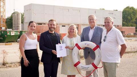 Frau Stolz steht mit vier weiteren Personen vor einer Baustelle - in den Händen halten sie einen Schwimmreifen und einen Förderbescheid.